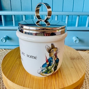 May include: A white ceramic egg cup with a silver-colored lid and handle. The cup features a colorful illustration of a rabbit in a blue jacket. The words "ate some" are printed on the side. The egg cup is on a wooden stand.