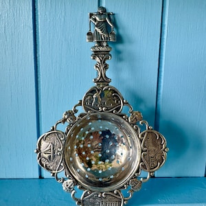 May include: An antique silver tea strainer with a decorative handle. The strainer has a round bowl with small holes and is surrounded by detailed embellishments, including architectural and nautical motifs. The handle features a figure carrying buckets.