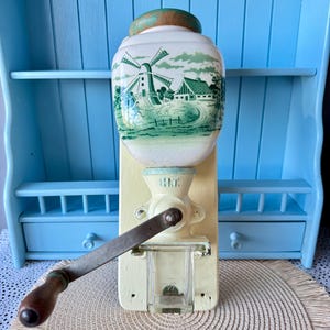 Op de afbeelding: Een vintage koffiemolen met een crèmekleurige behuizing en een keramische bovenkant met een groen-witte windmolenscène. Het heeft een metalen hendel en een houten knop. De molen staat tegen een lichtblauwe achtergrond.