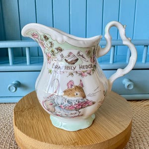 Puede incluir: Jarra de cerámica blanca con asa decorativa y base verde claro. La jarra presenta una ilustración de ratón y las palabras "Brambly Hedge". Acentos florales y de hojas adornan el borde.