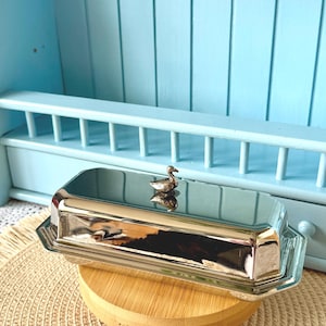 Könnte beinhalten: Eine silberfarbene Butterdose mit einem Entengriff. Die rechteckige Schale steht auf einem kleinen, runden Holzständer. Der Hintergrund zeigt ein hellblaues Regal und eine Wand.