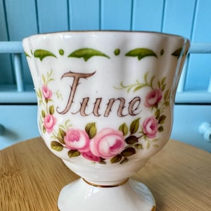 May include: A white porcelain egg cup featuring a fluted rim with a gold-tone edge. The front displays the word June in brown script, surrounded by hand-painted pink roses with green leaves and a decorative green leaf border along the top.