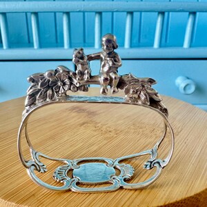 Peut inclure: Un rond de serviette en métal argenté présentant un motif sculpté d'un petit personnage assis sur une branche à côté d'un écureuil, entouré de motifs floraux et de feuilles en relief, avec une base ajourée et un centre ovale lisse.
