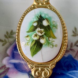 May include: A gold-toned spoon handle with a white porcelain oval featuring a green and white lily of the valley floral design.