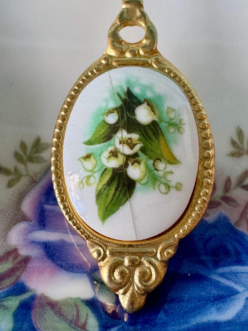 May include: A gold-toned spoon handle with a white porcelain oval featuring a green and white lily of the valley floral design.