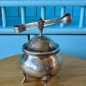Op de afbeelding: Een kleine, antieke zilveren suikerpot met een deksel en bijpassende tang. De kom heeft drie sierlijke voetjes en een decoratieve handgreep op het deksel. De tang rust bovenop de kom. De achtergrond is lichtblauw.