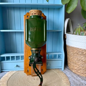 Könnte beinhalten: Eine Wandkaffeemühle mit einem dunkelgrünen Keramikbehälter, auf dessen Vorderseite das Wort KOFFIE eingeprägt ist. Die Mühle ist auf einem rechteckigen Holzbrett montiert und verfügt über eine Metallkurbel mit einem Holzgriff am Ende.