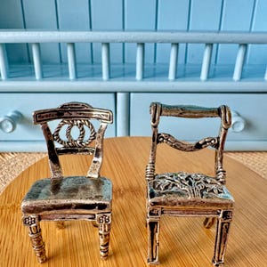 May include: Two miniature silver chairs with intricate details. One chair features a rope design on the back, while the other has a floral pattern on the seat. Both chairs have ornate legs and are displayed on a wooden surface.