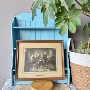 Könnte beinhalten: Ein gerahmtes Schwarz-Weiß-Familienporträt steht auf einem kleinen Holzständer vor einem hellblauen Regal. Das Porträt befindet sich in einem braunen Rahmen. Rechts befindet sich ein geflochtener Korb mit weißem Deckel und einer grünen Pflanze.