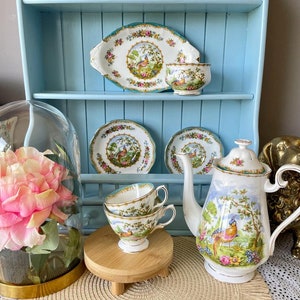 Puede incluir: Un juego de té vintage con un diseño de faisán y flores. El juego incluye una tetera, un azucarero, una jarra de crema, dos tazas y platillos, y una fuente para servir. El juego se exhibe en un estante azul con una base de madera.