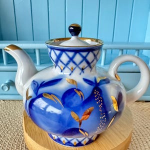 Könnte beinhalten: Eine weiß-blaue Porzellan-Teekanne mit goldenem Rand und Akzenten. Die Teekanne hat ein blaues Blumendesign und einen passenden Deckel mit einem dunkelblauen Knopf. Die Teekanne steht auf einem kleinen Holzständer.