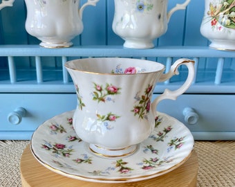 Può includere: Una tazzina da tè e un piattino bianchi con un motivo floreale. La tazza e il piattino sono su un supporto in legno. La tazza ha un bordo dorato e un delicato disegno floreale con fiori rosa e blu. Il piattino ha un design simile.