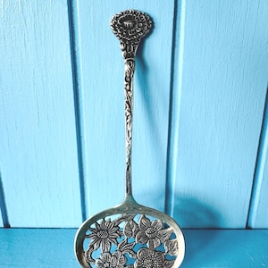 May include: A silver-toned serving spoon with a floral design. The spoon features an oval-shaped head with cut-out flowers and a long, slender handle. The handle is adorned with intricate floral patterns, and the top has a flower-shaped design.