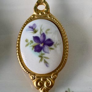 May include: A gold-toned spoon with a white porcelain handle featuring a purple flower with green leaves. The handle is oval-shaped with a decorative border.