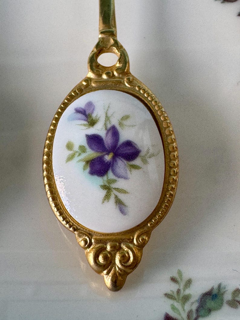 May include: A gold-toned spoon with a white porcelain handle featuring a purple flower with green leaves. The handle is oval-shaped with a decorative border.