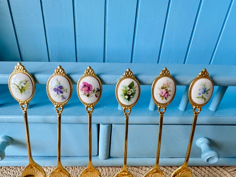 May include: Six gold teaspoons with white porcelain handles decorated with floral designs. Each spoon features a different floral pattern, including pink roses, purple flowers, and white lilies.