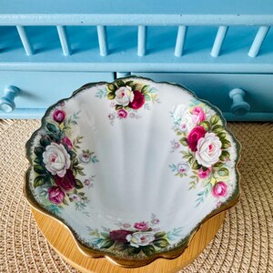 Può includere: Un piatto bianco con bordo smerlato con un motivo floreale di rose nei toni del rosa, rosso e bianco, accentuato da foglie verdi. Il piatto ha un bordo dorato e poggia su una base di legno rotonda. Lo sfondo è una mensola azzurra.