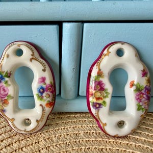 May include: Two oval porcelain keyhole escutcheons featuring a white base with colorful floral patterns, including pink roses and small blue and yellow blossoms. The edges are trimmed in deep magenta and accented with delicate gold filigree details.