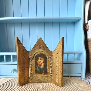 Pode incluir: Um tríptico de cor dourada com um painel central arqueado contendo uma pintura de uma figura segurando uma criança, ladeado por dois painéis laterais articulados. A moldura é decorada com padrões em relevo intrincados, detalhes vermelhos e topos pontiagudos.