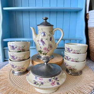Könnte beinhalten: Ein cremefarbenes Teeservice bestehend aus einer Teekanne, einer Zuckerdose mit Deckel und sechs kleinen Schalen. Jedes Teil ist mit einem Blumenmuster aus violetten und rosa Blüten verziert. Teekanne und Zuckerdose haben zinngraue Deckel und Füße.