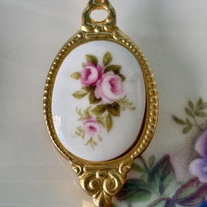 May include: A gold-toned spoon with a white porcelain oval handle featuring a pink rose floral design.