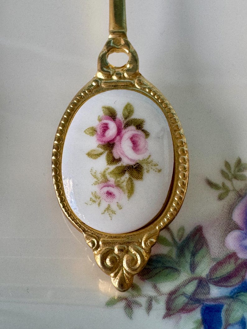 May include: A gold-toned spoon with a white porcelain oval handle featuring a pink rose floral design.