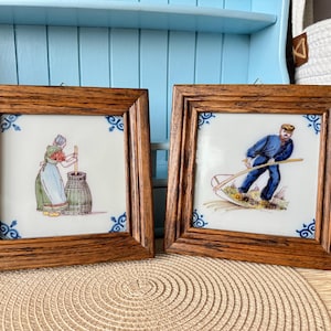 May include: Two framed ceramic tiles with brown wooden frames. One tile depicts a person churning butter, the other shows a person working in a field. Both tiles have a white background with blue corner designs.