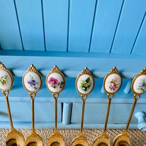 May include: Six gold-tone teaspoons with white porcelain handles featuring floral designs. Each spoon has a different floral pattern.