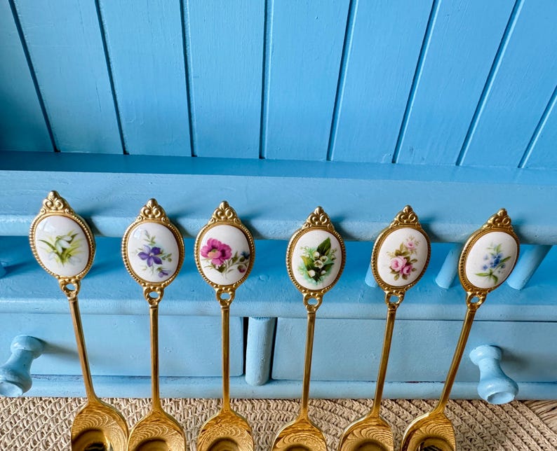 May include: Six gold-tone teaspoons with white porcelain handles featuring floral designs. Each spoon has a different floral pattern.