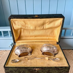 May include: An antique silver salt cellar set with two ornate bowls and two small spoons, presented in a green and gold velvet-lined box. The silver bowls have detailed relief patterns.