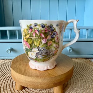 May include: A white ceramic mug with a scalloped base and a decorative handle, featuring a colorful illustration of blackberries, pink flowers, and a small figure in a yellow hat nestled among green foliage and a stone archway.