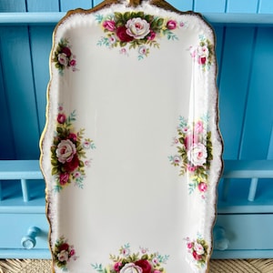 Puede incluir: Una bandeja rectangular de cerámica blanca con un borde dorado festoneado. La bandeja está decorada con racimos de rosas rosas y rojas y hojas verdes. Una pieza decorativa para servir alimentos o exhibir artículos.