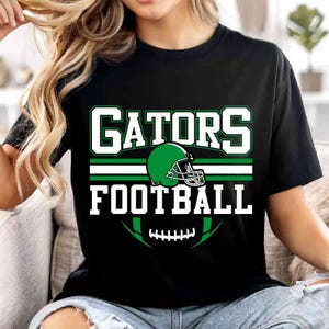 Peut inclure: Un t-shirt noir à manches courtes avec une impression graphique portant le texte GATORS FOOTBALL en lettres capitales blanches. Le motif comprend un casque de football américain vert, une bande horizontale blanche et verte et un ballon vert.