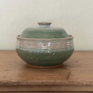 May include: A handmade ceramic lidded bowl in shades of green and beige. The bowl features a textured floral pattern around the base and a matching lid with a small knob. The piece is set on a wooden surface, showcasing its rustic charm.