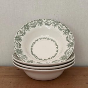 Może przedstawiać: Stos czterech białych ceramicznych misek z zielonym kwiatowym wzorem na obrzeżach. Miski są ułożone jedna na drugiej. Miski stoją na drewnianej powierzchni. Średnica około 15 cm.