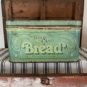 Peut inclure: Boîte à pain vintage en métal de couleur turquoise avec l'inscription "Wheat Heart Bread" dans une police décorative. La boîte a un couvercle à charnière et est ornée d'éléments floraux. Le texte "The Finest Quality Since 1843" est également visible.