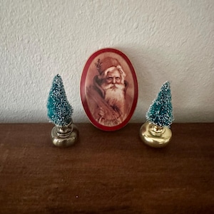 Könnte beinhalten: Eine weihnachtliche Dekoration mit einem ovalen Santa Claus-Bild mit rotem Rand, flankiert von zwei kleinen, teal-farbenen Flaschenbürstenbäumen auf goldenen Sockeln. Die Elemente sind auf einer dunklen Holzoberfläche angeordnet.