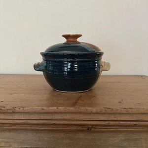 Puede incluir: Una cazuela de cerámica de color verde azulado oscuro con tapa y dos asas pequeñas. La tapa tiene un pomo marrón. El plato está sobre una superficie de madera.