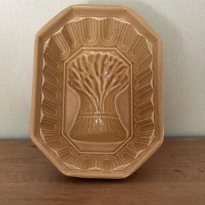 May include: Tan ceramic mould with a wheat sheaf design. The rectangular mould has an octagonal shape with a scalloped border. The wheat sheaf is in the centre of the mould. The mould is on a wooden surface.