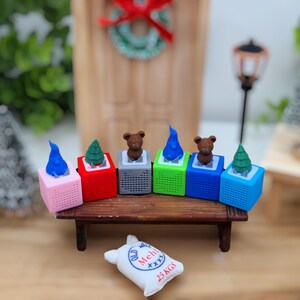 Könnte beinhalten: Sechs Miniatur-Weihnachtslautsprecher in verschiedenen Farben, darunter rosa, rot, grau, grün, blau und hellblau, mit einem kleinen Bären, einem Weihnachtsbaum und einer blauen Flamme auf jedem Lautsprecher. Die Lautsprecher sind in einer Reihe auf einem Holzregal angeordnet. Ein kleiner weißer Sack mit dem Text "Old Mehl 25 KGS" befindet sich im Vordergrund.