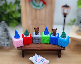 Può includere: Sei mini altoparlanti a tema natalizio in vari colori, tra cui rosa, rosso, grigio, verde, blu e blu chiaro, con un piccolo orso, un albero di Natale e una fiamma blu sulla parte superiore di ogni altoparlante. Gli altoparlanti sono disposti in fila su uno scaffale di legno. Un piccolo sacchetto bianco con il testo "Old Mehl 25 KGS" è in primo piano.