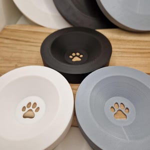 Può includere: Quattro ciotole per animali domestici in ceramica di diversi colori, ciascuna con un design a forma di impronta di zampa. Le ciotole sono impilate, con la più grande in basso e la più piccola in alto. Le ciotole sono bianche, nere, grigie e grigio chiaro.