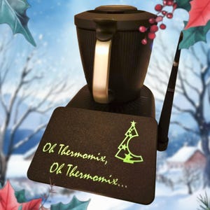 Peut inclure: Un appareil de cuisine noir avec une poignée métallique et un tapis rectangulaire noir orné d'un motif vert clair représentant un sapin de Noël stylisé et le texte "Oh Thermomix, Oh Thermomix..." écrit en police cursive vert clair.
