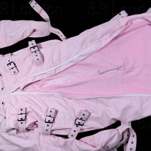 Può includere: Un indumento rosa chiaro a maniche lunghe con collo alto, cerniera centrale bianca e molteplici cinghie regolabili con fibbie nere sul petto e sulle maniche. Il capo presenta cuciture rinforzate e occhielli metallici sulle cinghie.