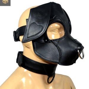 Puede incluir: Máscara de perro de cuero negro con orejas grandes y un aro en la nariz. La máscara cubre los ojos y la nariz, con un collar a juego. Las orejas tienen detalles en forma de corazón. El collar tiene un anillo de metal.