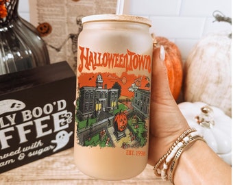 May include: A brown and white reusable tumbler with a Halloween town design. The tumbler has a black lid and a straw. The design features a spooky house, a pumpkin, and the text "Halloween Town Est. 1998".