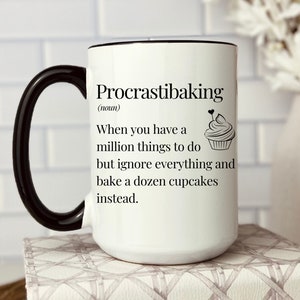 May include: White ceramic mug with a black rim and handle. The mug has a black graphic that says "Procrastibaking (noun) When you have a million things to do but ignore everything and bake a dozen cupcakes instead." with a small cupcake illustration.