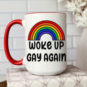 May include: White ceramic coffee mug with a red rim and handle. The mug features a rainbow graphic and the text "Woke Up Gay Again".