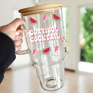 May include: A clear glass tumbler with a bamboo lid and a straw. The tumbler has a design of watermelons and the text "Cortisol Cocktail" in pink and white.