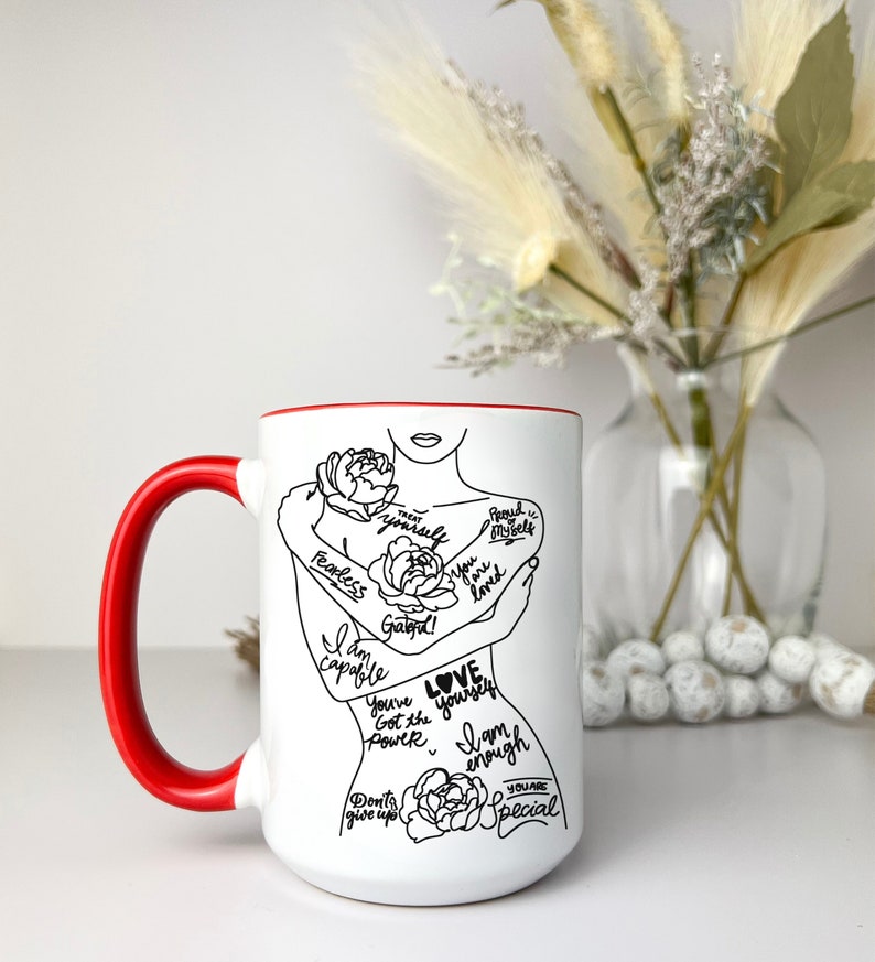 Puede incluir: Taza de cer&aacute;mica blanca con borde y asa rojos. La taza presenta un dibujo en l&iacute;nea negra de una mujer abraz&aacute;ndose a s&iacute; misma con las palabras "Love yourself" y otras afirmaciones positivas.